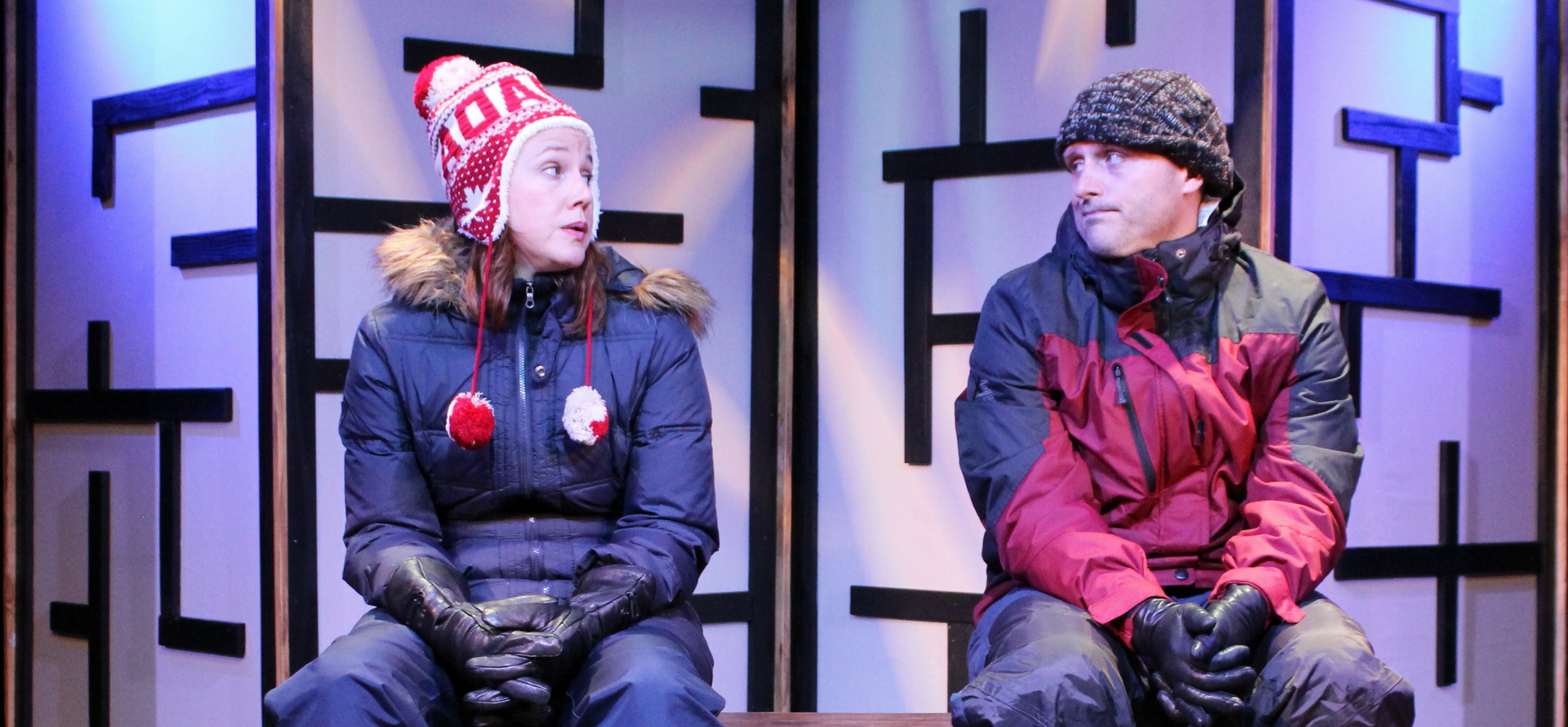 A man and woman on stage in play, sitting on chairs wearing snow gear